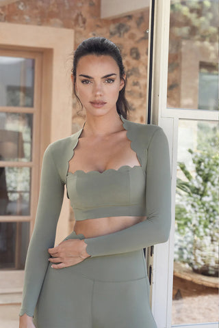 A woman with dark hair styled in a high ponytail stands near a glass door, wearing the Serene Stretch: All-Hours Long Sleeve Top in Sage. The top is paired with matching high-waisted bottoms, both featuring scalloped edges. A blurred outdoor scene and part of a stone-walled building are visible in the background.