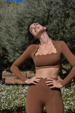 A woman stands outdoors, wearing the Serene Stretch: All-Hours Long Sleeve Top paired with scalloped-edged leggings. She has her hands on her hips and is looking up, eyes closed, with a joyful expression. The background features greenery, including trees and white flowers.