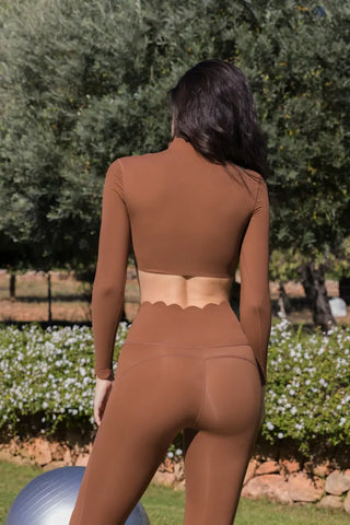 A woman with long dark hair stands outdoors with her back to the camera, wearing the Serene Stretch: All-Hours Long Sleeve Top and matching leggings. The background features greenery, including trees and bushes, with a pathway lined by stones and flowers. An exercise ball is partially visible.
