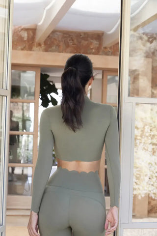 A woman with long dark hair in a ponytail is seen from behind, walking indoors. She is wearing a form-fitting, olive-green workout outfit consisting of the Serene Stretch: All-Hours Long Sleeve Top with scalloped edges and matching high-waisted leggings. The room has wooden and glass-paneled doors and light brown walls.