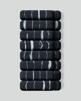 A stack of ten black cylindrical pads, with white marbled streaks, is neatly aligned in two vertical rows against a pale gray background. The top pad features an embossed logo. Product: Vibrant Bangles Set for an Energizing Look.