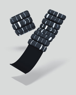 The Vibrant Bangles Set in Stone features black rectangular modules with white stripes arranged vertically. One column stands upright, another is slightly curved, and a flat black strip extends horizontally from their base against a plain light gray background with visible shadows below.