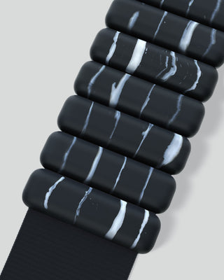 The Vibrant Bangles Set features black, rectangular stones with white wavy streaks, arranged in rows and connected by a dark strap with a textured pattern against a soft, light gray background.