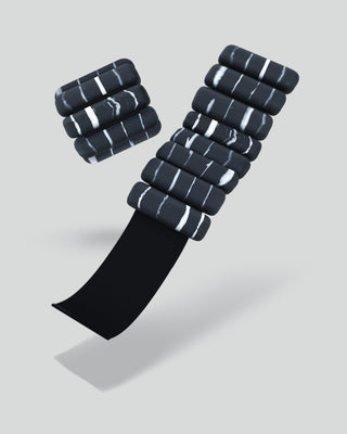 Black, pill-shaped Vibrant Bangles with white marbling are elegantly stacked on a curved black surface. A light gray background creates a striking contrast, enhancing their energizing look and casting soft shadows.