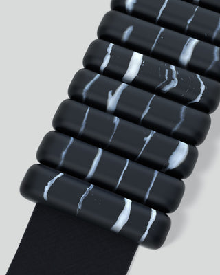 Close-up of a modern black bangle from the Vibrant Bangles Set for an Energizing Look. It features cylindrical beads with irregular white streaks, creating a marbled effect, laid diagonally on a light gray background to highlight its sleek and contemporary design.