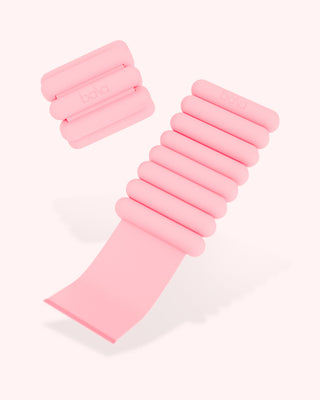 Two pink weights from the Vibrant Bangles Set for an Energizing Look are on a light background. One is extended, showing its adjustable strap; the other is folded, displaying its compact design. They have cylindrical segments for even weight distribution and feature "bala" embossed on them.
