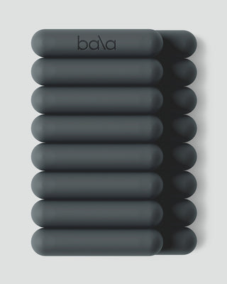 A set of ten dark gray circular Vibrant Bangles, shown in two columns of five on a light gray background, with the top bangle featuring the brand name "bala" in lowercase black letters. Each bangle is smooth and cylindrical with rounded ends.