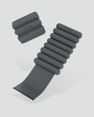 Two black weighted wristbands by "bala" are displayed, one unsnapped to reveal its elastic strap. The bands feature horizontal cylindrical weights and combine style with functionality for fitness. The set is called Vibrant Bangles Set for an Energizing Look, with a light gray background.
