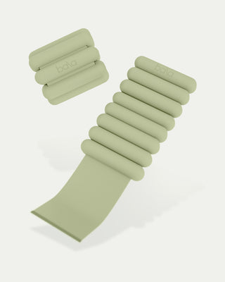 The Vibrant Bangles Set for an Energizing Look features two sage green exercise weights. One, a wrist weight with fastened strap and cushioned segments, and the other with an extended strap. Both have "bala" branding and rest on a neutral background.