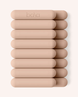 Ten beige cylindrical Vibrant Bangles are stacked vertically on a light background. The top bangle has "bala" embossed on it. Each piece has a soft matte finish and rounded edges, creating a clean and minimalist arrangement.
