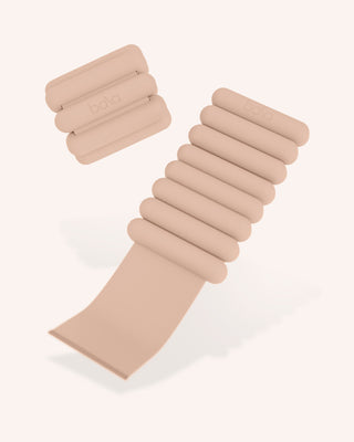 The Vibrant Bangles Set for an Energizing Look features a beige resistance band set with a modern design, including a longer band with evenly spaced cylindrical segments and a shorter band of three segments. Both embossed with a logo, they are showcased against a plain, light background.