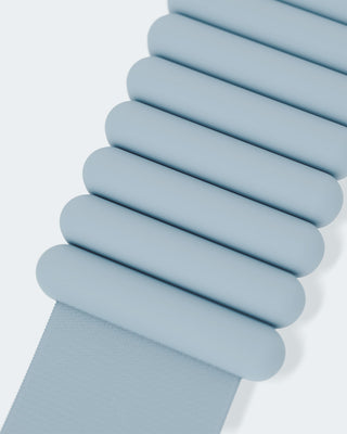 A close-up features a row of light blue cylindrical Vibrant Bangles connected and horizontally aligned on a flat surface, attached to a fabric strap that extends out of frame at the bottom against a light, neutral background.