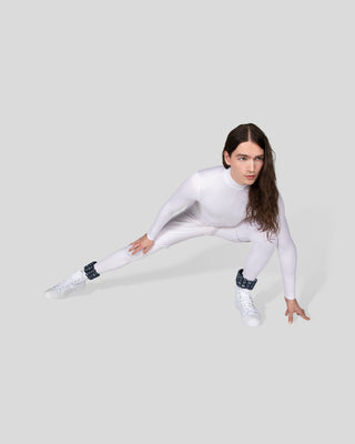 A person with long brown hair strikes an athletic pose in a fitted white bodysuit and sneakers. One leg bent, hand on the floor, they exude focus against a light gray background. Complement their style with a Vibrant Bangles Set for an Energizing Look.