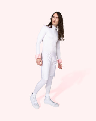 A person with long hair stands against a light pink background, wearing a fitted white outfit with pink ruffled cuffs, white shorts, and white high-top sneakers. They accessorize with the Vibrant Bangles Set for an Energizing Look while gazing left in a relaxed posture.