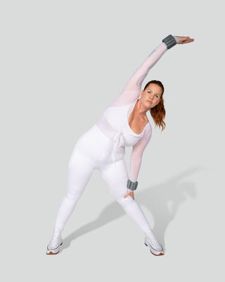 In a plain, light gray setting, a person in a white workout outfit and sneakers performs a side stretch using the Vibrant Bangles Set for an Energizing Look. They lean right with one arm raised overhead while the other touches their knee, displaying a focused expression.