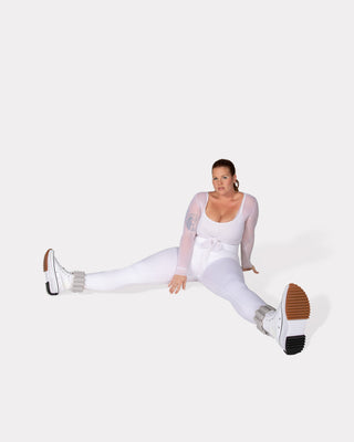 A person wearing a sheer white outfit and chunky white boots with brown soles demonstrates flexibility in a floor split. They showcase tattoos, hoop earrings, and vibrant bangles for an energizing look, with their hair pulled back against a plain white backdrop.