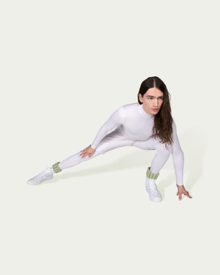 A person with long hair, dressed in a white, form-fitting outfit and white sneakers, stretches in a lunge on a light background. Green ankle cuffs from the Vibrant Bangles Set for an Energizing Look adorn each ankle over their shoes as their left hand touches the ground.