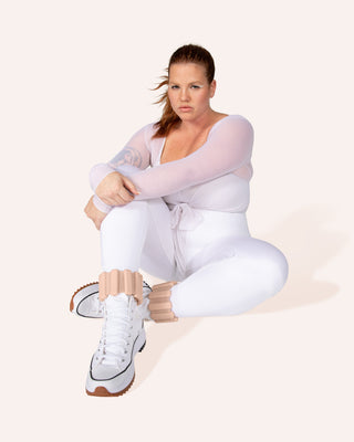 A person with red hair in a ponytail sits on the floor wearing a sheer white bodysuit and leggings, paired with white high-top sneakers featuring beige ankle cuffs. The background is off-white. They look directly at the camera with a neutral expression, embodying the essence of the Vibrant Bangles Set.