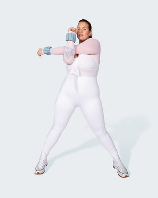 A person in a fitted white outfit and white sneakers stands on a light background, stretching their left arm across their chest with blue wrist weights. They have tattoos on their left arm, wear hoop earrings, and accessorize with the Vibrant Bangles Set for an Energizing Look.