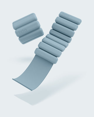 A pair of gray-blue adjustable wrist or ankle weights from the "Vibrant Bangles Set for an Energizing Look" features the brand name "bala" engraved on each segment. Displayed on a light background, one strap is extended and the other curled, showcasing their soft, segmented design.