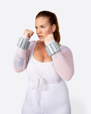 A woman in a sheer white outfit with front tie and Vibrant Bangles Set holds a guarded stance, fists near her face. Her hair is pulled back, tattoo on her left forearm, she gazes intently against a plain white background.