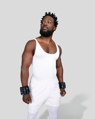 A man with a beard and short, textured hair stands seriously against a gray backdrop, wearing a white sleeveless leotard, shorts, and the Vibrant Bangles Set for an Energizing Look on his wrists.