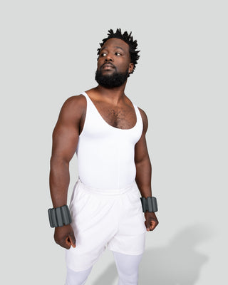 Against a plain background, a man exudes confidence wearing a white tank top and pants. His short twists add style to his black hair, while dark wrist weights complement the strength of his serious expression and upright stance.