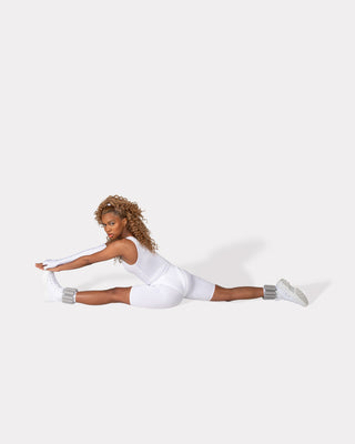 A person with long, curly hair stretches in a split position on a white background. Wearing all-white athletic gear and ankle weights, they extend their arms toward their left foot. Shadows enhance the scene's vibrant energy, perfectly paired with the Vibrant Bangles Set for an Energizing Look.