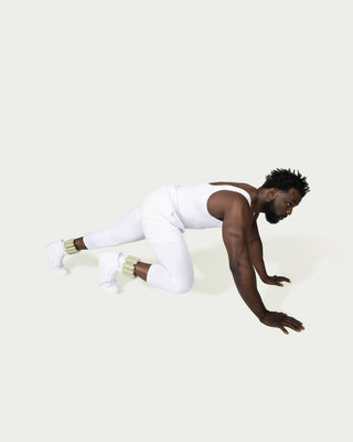 Wearing white athletic clothing and light green leg warmers, the person is mid-crawl, with hands and one knee on the ground and the other knee raised, against a plain light gray backdrop.