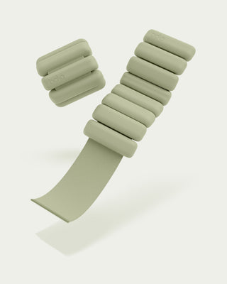The Vibrant Bangles Set for an Energizing Look includes sleek, minimalist sage-colored bangles. Each bangle has cylindrical weights on a strap with one open to show the strap's texture. The word "bala" is embossed on the weights.