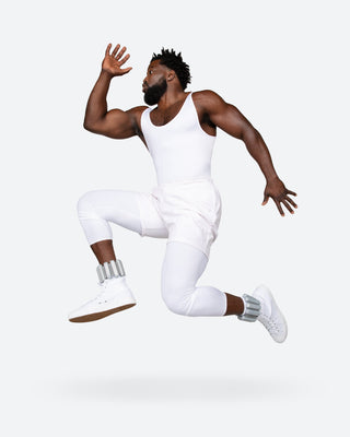 A man jumps mid-air against a light background, wearing a white tank top, shorts, leggings, ankle weights, and high-top sneakers. His right arm is bent upward and his left extended back. He sports an accessory from the Vibrant Bangles Set for an Energizing Look.