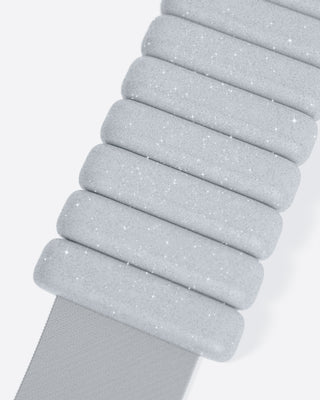 A close-up image of a light gray, quilted strap exhibits padded segments with a sparkly texture and star-like specks, giving it a shimmering appearance.