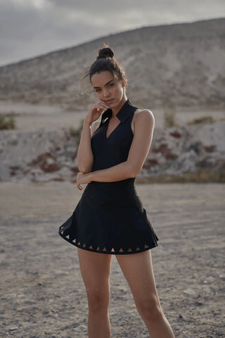 A woman stands confidently in an arid, rocky landscape, wearing the AeroPolo™ Sleeveless Active Performance Tank Top with a flared skirt featuring triangular cutouts. Her hair is styled in a bun, and she gazes directly at the camera with one hand near her chin. The overcast sky provides a soft lighting effect.