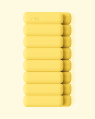 A stack of nine soft yellow blocks from the Vibrant Bangles Set creates a harmonious and minimalistic look on a light yellow background. The top block subtly shows the Bala logo, while each piece has rounded edges and a smooth texture, arranged vertically.