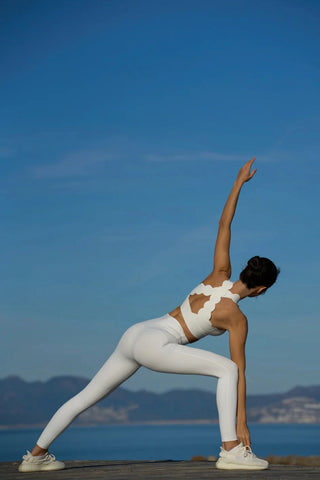 A person wearing the PeaceActive™ - CrissCross Performance Sports Bra stretches against a backdrop of a clear blue sky and distant mountains. They are bending sideways with one arm reaching up and the other touching their ankle, standing on a rocky surface with a calm ocean in the background.