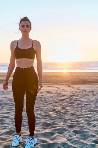 A woman stands on a sandy beach during sunset. She is dressed in black athletic wear, including a scalloped-edge sports bra and the 7/8 High-Waist Energy Legging. She has white sneakers on her feet. The sun is low on the horizon, casting a warm glow over the ocean and beach.