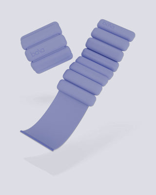 Two lilac Vibrant Bangles from the Energizing Look set float against a light gray background. One bangle is stretched, showcasing its flexibility and rounded weights, while the other lies stacked with the "bala" logo visible, highlighting their compact design.