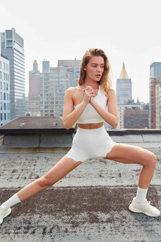 A person with long hair is doing a stretching exercise on a rooftop. They are dressed in an Elegant Zen One Shoulder Activewear Bra, high-waisted shorts, and white sneakers with white socks. The background features multiple high-rise buildings under a cloudy sky. The person appears focused and balanced.