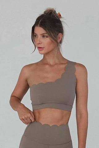 A woman stands against a plain light grey background wearing the Elegant Zen One Shoulder Activewear Bra in khaki brown, paired with matching high-waisted leggings. Her hair is tied up in a loose bun with a brown scrunchie, and she has a composed expression while looking slightly to her left.