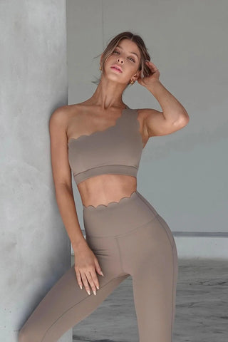 A woman stands against a gray wall, confidently posing with one hand resting on her head and gazing forward. She is wearing a matching taupe workout set that includes the Elegant Zen One Shoulder Activewear Bra, which features an asymmetrical design and scalloped edges, paired with high-waisted leggings adorned with similar scalloped trim.