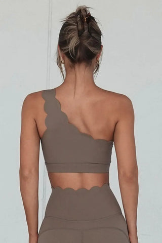 A woman with an updo hairstyle is seen from behind wearing the Elegant Zen One Shoulder Activewear Bra, a taupe-colored, asymmetrical sports bra featuring a scalloped edge on the single strap and waistband, paired with matching high-waisted leggings that also feature a scalloped edge at the top.