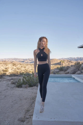 A woman wearing the CrissCross Performance Sports Bra from PeaceActive™ stands on a concrete ledge near a pool, nestled within a desert landscape. She gazes off to the side, with a mountain range and clear blue sky forming the serene backdrop, all softly illuminated by daylight.