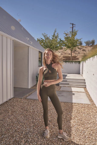 A woman with long blonde hair is posing outdoors in front of a modern white building. She is wearing the PeaceActive™ CrissCross Performance Sports Bra, a green sleeveless athletic outfit, and beige sneakers. The background features a clear blue sky, trees, gravel, and rectangular stepping stones leading to the entrance.