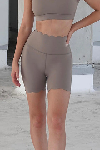 A person is shown from the neck down, wearing a light brown workout outfit that includes a sleeveless crop top and the FitFlex - High-Waist Mini Workout Shorts, which feature a unique scalloped edge at the waist and thigh hems. The background features unfinished concrete walls and flooring.