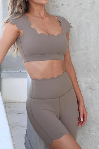 A person with long, wavy hair wears a beige scalloped-edge crop top paired with FitFlex High-Waist Mini Workout Shorts. The outfit features a wavy, scalloped design along the edges. They are standing against a light gray concrete wall and a large window, with partial natural light illuminating the scene.