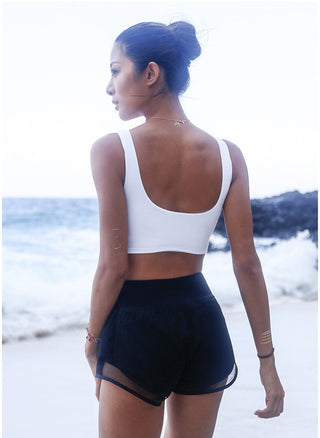A woman stands on a beach with her back to the camera, wearing a Scoop Back Cover Tank in white along with black shorts. Her hair is styled in a bun, and she adorns temporary metallic tattoos on her arms. The ocean serves as the backdrop, while an overcast sky looms above and waves gently lap against the shore.