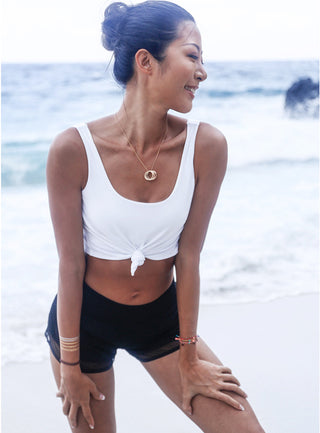A woman stands on the beach, smiling and looking to the side. She wears a white Scoop Back Cover Tank that ties at the front and black shorts. Her hair is styled in a bun, and she has several bracelets adorning her wrists. The ocean waves gently crash in the background under a bright, slightly overcast sky.