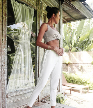 A woman in a casual outfit stands on a wooden porch, gazing to the side. She wears the Scoop Back Cover Tank and white pants. Behind her, there is a glass door with curtains and lush greenery, including large banana leaves and a sunbed, creating a serene outdoor setting.