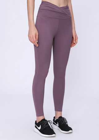 A person is wearing PE High Rise Tight Pants in mauve featuring a crossover waistband, paired with black and white athletic shoes, against a plain white backdrop. The image highlights the fit and fabric texture on their legs and waist area.