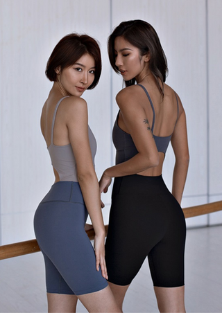 Two women stand by a ballet barre against a white wall, both wearing the Simply Sexy Unitard. One unitard is in gray and navy, the other in gray and black. With short hair styled neatly, they show side profiles while appearing focused on their practice.